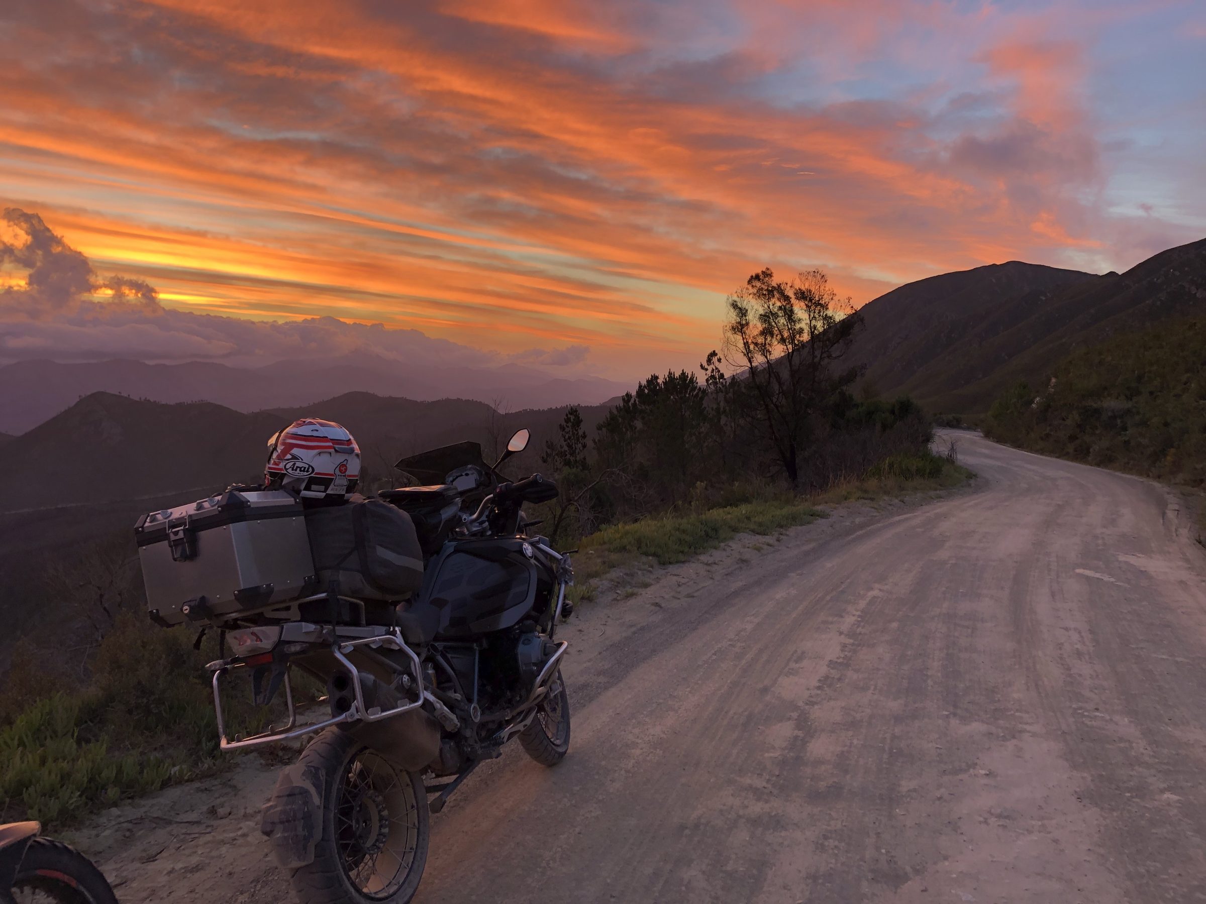 A sunset over the mountains, gravel road winding ahead, motorcycle in the foreground